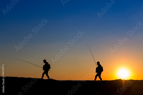 3 Fishermen silhouette with upright rods stroll walk on the rocks at dawn dusk sunrise sunset. The sky is blue purple orange. They have upright rodsat dawn 