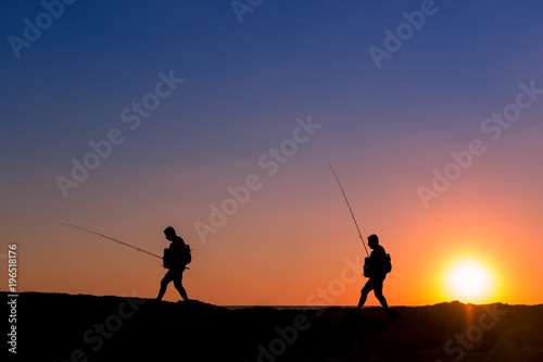 3 Fishermen silhouette with upright rods stroll walk on the rocks at dawn dusk sunrise sunset. The sky is blue purple orange.  They have upright rodsat dawn   © dan