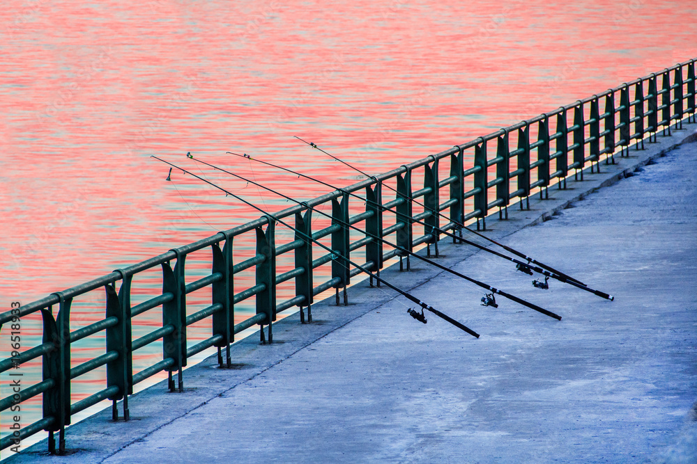 Four fishing-rods without people stand on the bank of the river, leaning on  the fence. Stock Photo