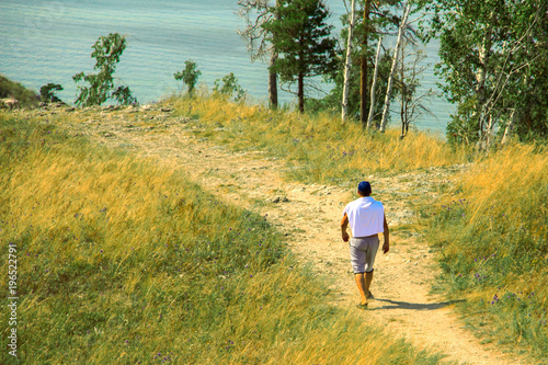 A man in shorts  a white T-shirt and a blue cap walking along a sandy and stony path among bushes  grass and flowers.