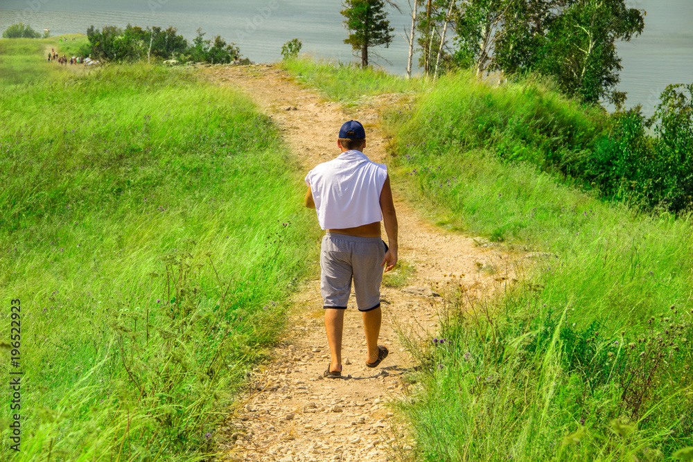A man in shorts, a white T-shirt and a blue cap walking along a sandy and stony path among bushes, grass and flowers. A man in shorts, a white T-shirt and a blue cap walking along a sandy and stony path among bushes, grass and flowers.