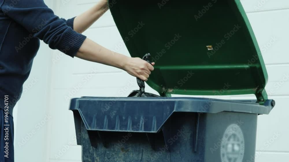 An unrecognizable woman throws a bag of garbage into a garbage can near the house