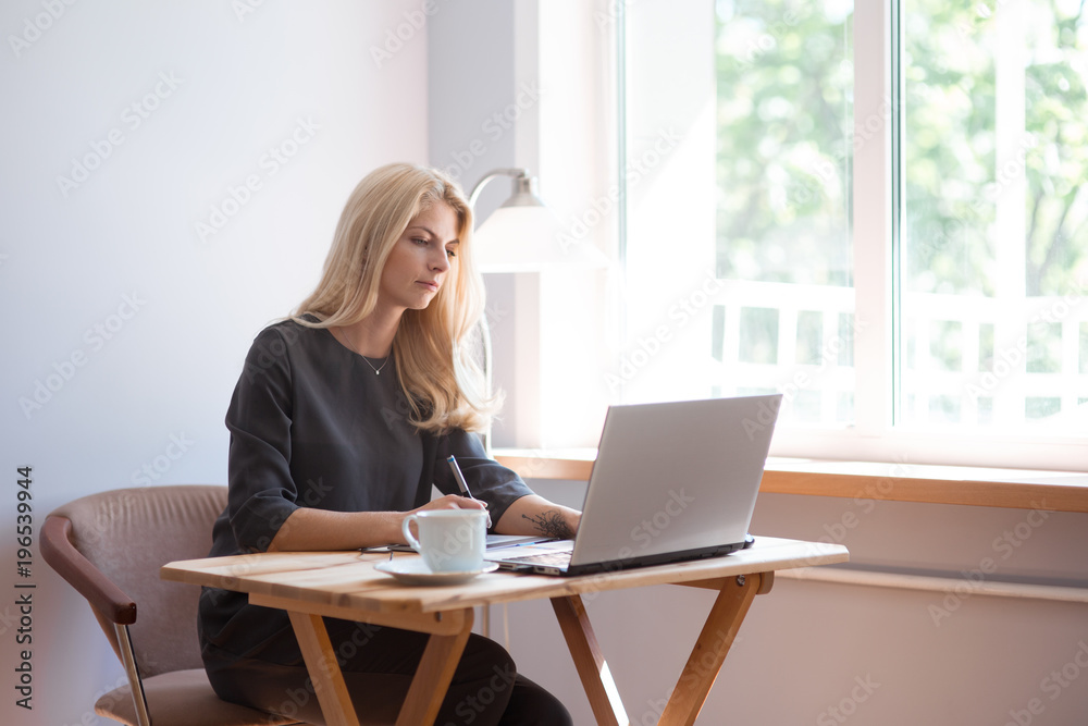 Young blonde caucasian woman photographer sitting at the laptop with graphic tablet wooden table large window and grey wall working in grey dress smiling drinking coffee and camera lens on surface