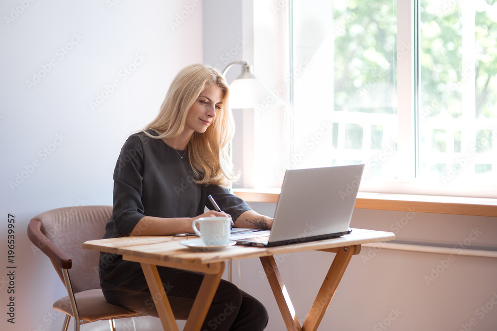 Young blonde caucasian woman photographer sitting at the laptop with graphic tablet wooden table large window and grey wall working in grey dress smiling drinking coffee and camera lens on surface
