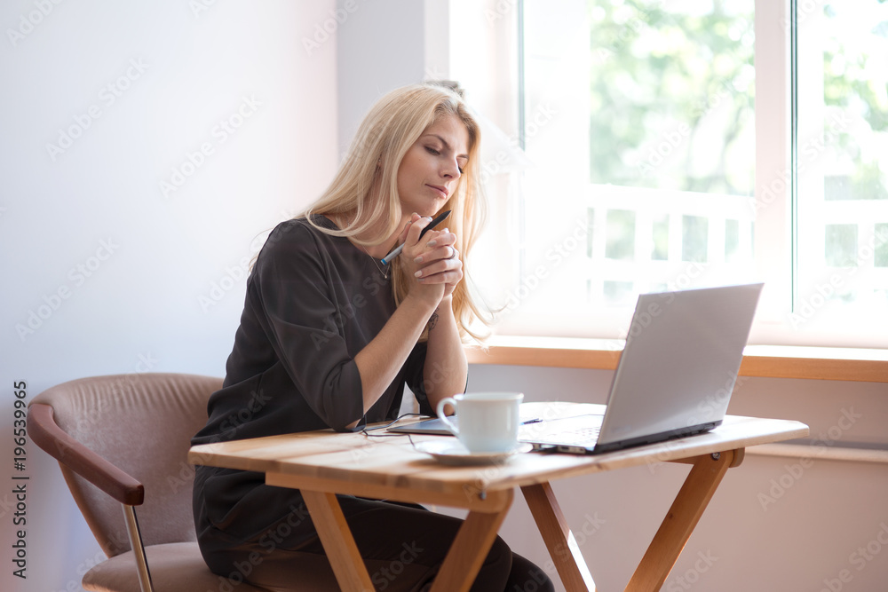 Young blonde caucasian woman photographer sitting at the laptop with graphic tablet wooden table large window and grey wall working in grey dress smiling drinking coffee and camera lens on surface