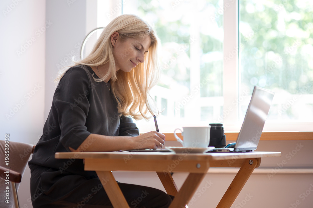 Young blonde caucasian woman photographer sitting at the laptop with graphic tablet wooden table large window and grey wall working in grey dress smiling drinking coffee and camera lens on surface Young blonde caucasian woman photographer sitting at the laptop with graphic tablet wooden table large window and grey wall working in grey dress smiling drinking coffee and camera lens on surface