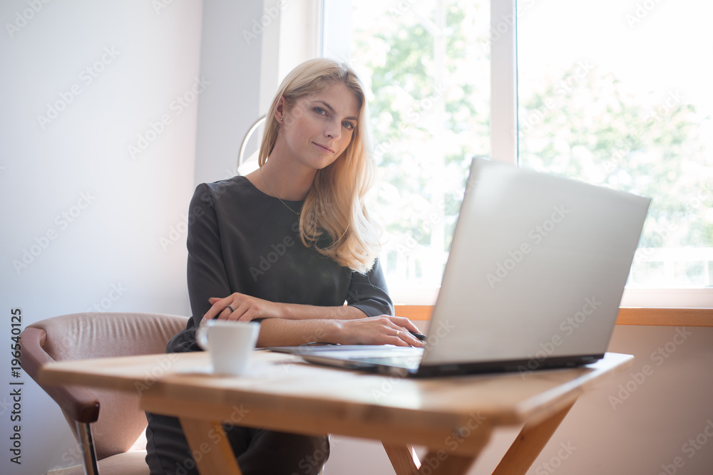 Young blonde caucasian woman photographer sitting at the laptop with graphic tablet wooden table large window and grey wall working in grey dress smiling drinking coffee and camera lens on surface Young blonde caucasian woman photographer sitting at the laptop with graphic tablet wooden table large window and grey wall working in grey dress smiling drinking coffee and camera lens on surface