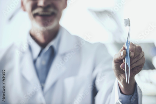 Portrait of a smiling senior dentist holding toothbrush on the background his patient in dental clinic