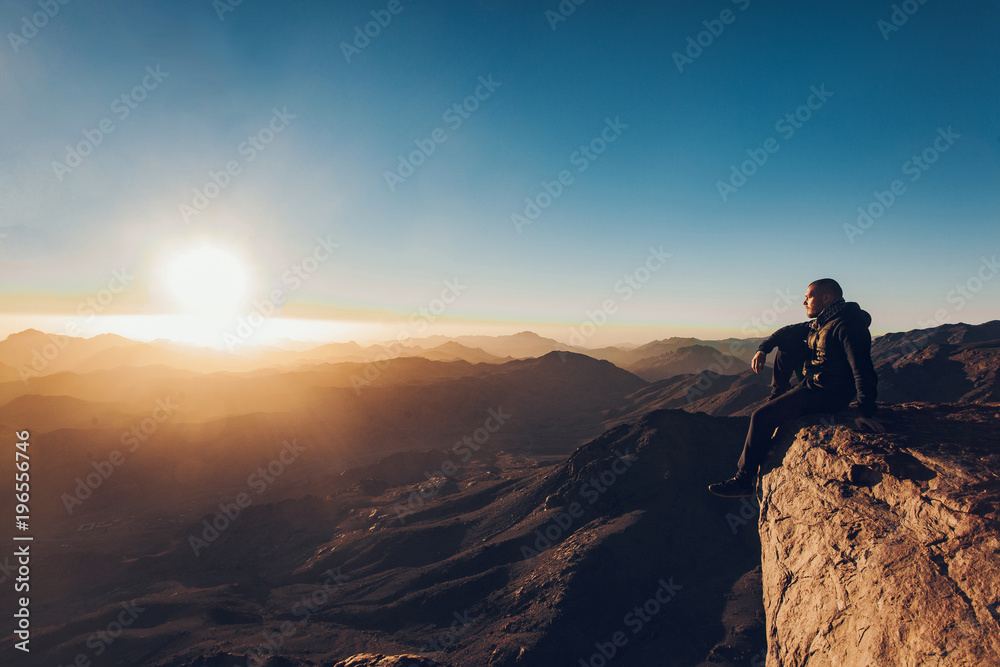 Fototapeta premium Man sits on edge of cliff on Mount Sinai against background of sunrise.