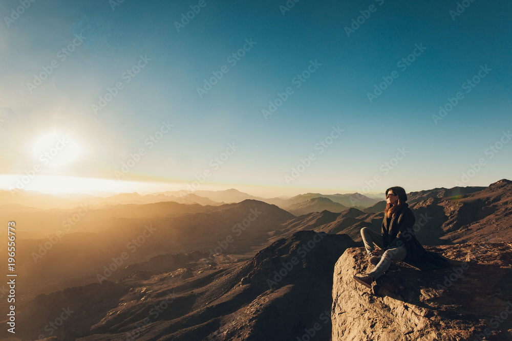 Woman sits on edge of cliff on Mount Sinai against background of sunrise.