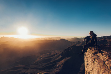 Man sits on edge of cliff on Mount Sinai against background of sunrise.