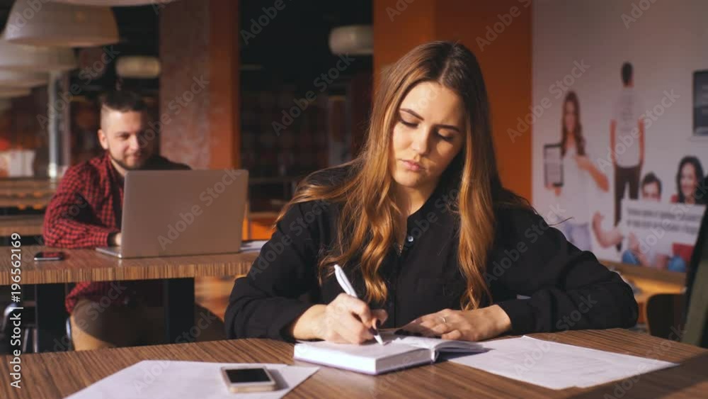 business young girl writing note in notebook in cafe, man with laptop on background business young girl writing note in notebook in cafe, man with laptop on background