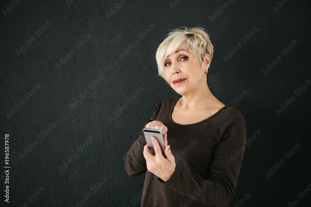 Fototapeta premium A positive modern elderly woman is holding a cell phone and is using it. The older generation and modern technology.