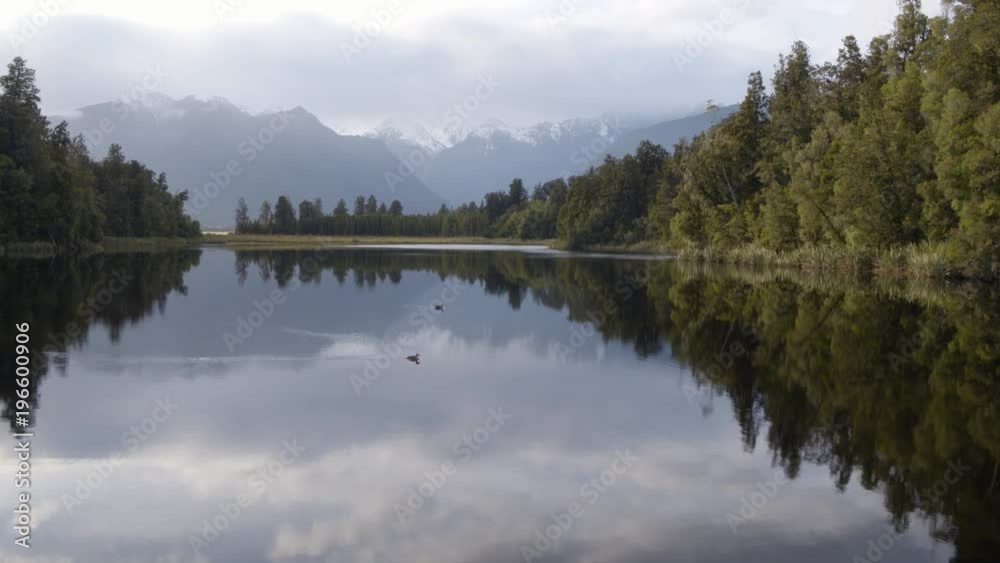 Drone shot pushing in on beautiful reflective lake with dark green trees and snowy mountains in the distance. Lake Matheson, West Coast, New Zealand. Drone shot pushing in on beautiful reflective lake with dark green trees and snowy mountains in the distance. Lake Matheson, West Coast, New Zealand.