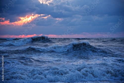 Stormy weather in the sea. Big waves incoming on the beach. Orange sun rays go through dark blue clouds.