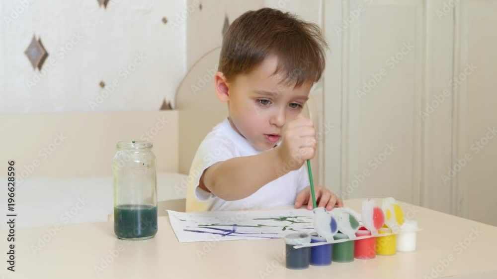 The child draws watercolor drawings on a white sheet of paper, sitting at the table. The child draws watercolor drawings on a white sheet of paper, sitting at the table.