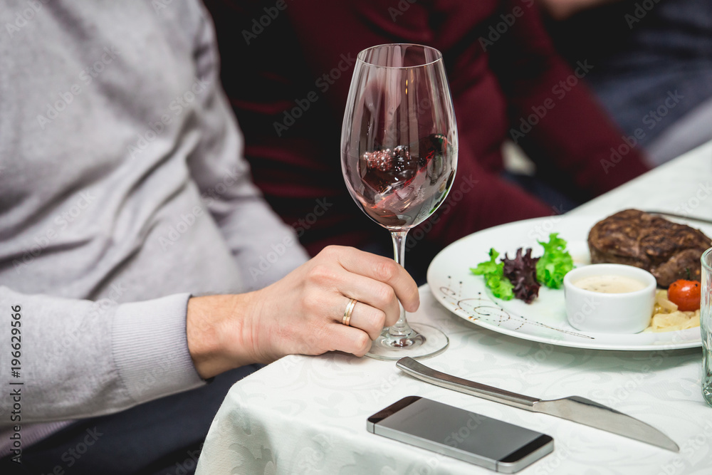 wine tasting in a restaurant. man holds glass of white wine. fresh grilled bbq roast beef steak and sauce on a white plate with green leaf of salad.