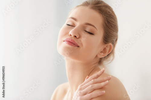Young woman applying cream onto her skin on light background