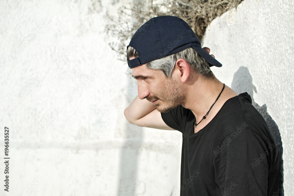 Urban, modern young man with black cap, in profile, looking straight ahead, next to a white wall of the street.