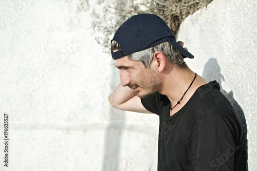 Urban, modern young man with black cap, in profile, looking straight ahead, next to a white wall of the street.