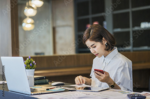 Beautiful woman using smartphone while working with laptop at modern office 