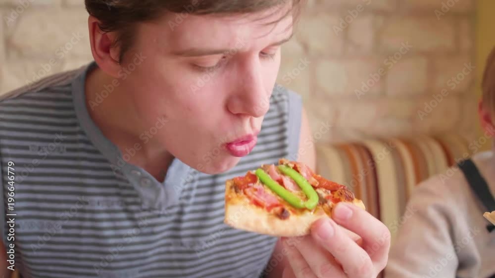 The guy takes and eats freshly baked pizza with cheese, ham, tomatoes and herbs . Italian cuisine The guy takes and eats freshly baked pizza with cheese, ham, tomatoes and herbs . Italian cuisine