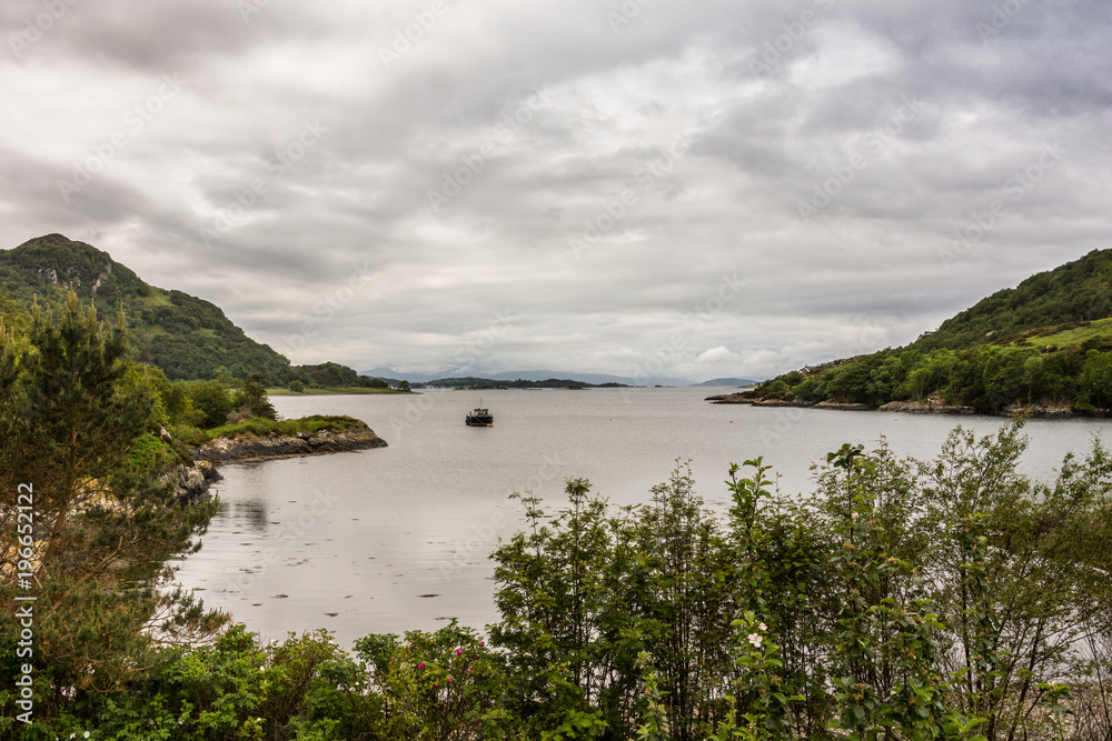 Stromeferry, Scotland - June 10, 2012: From Castle Strome ruins looking over Loch Carron towards ocean. Green hills on shores. Motor boat anchored in lake at distance. Rain-heavy cloudscape.