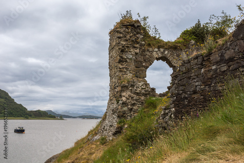Stromeferry, Scotland - June 10, 2012: Window in rock wall of Castle Strome ruins on green hill. Cloudscape and motor boat anchored on Loch Carron. Mountains on opposite shore and horizon.
