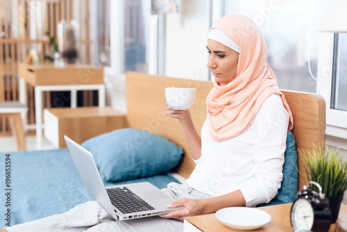 An arabian woman is drinking tea and working on the computer while sitting on the bed.