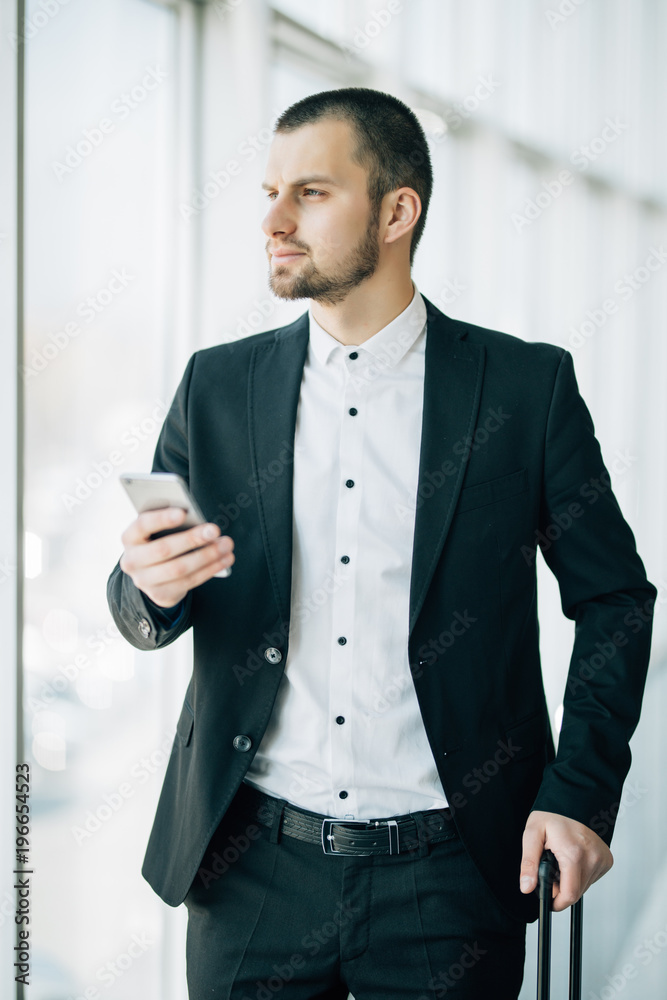 Young businessman with suitcase and using smart phone waiting for flight. Caucasian businessman waiting at airport terminal holding mobile phone. Young businessman with suitcase and using smart phone waiting for flight. Caucasian businessman waiting at airport terminal holding mobile phone.