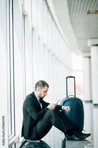 Young business man sitting on the phone with the suitcase at the airport waiting for the delayed flight