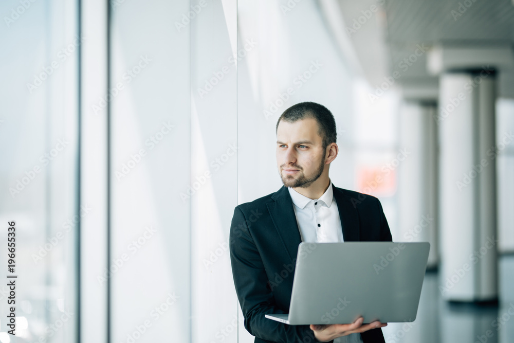 Businessman stands in a bright airy office holding an open laptop in his hands Businessman stands in a bright airy office holding an open laptop in his hands