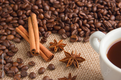 roasted coffee beans are scattered on beige burlap with cinnamon sticks and anise on the right in the corner a cup of espresso