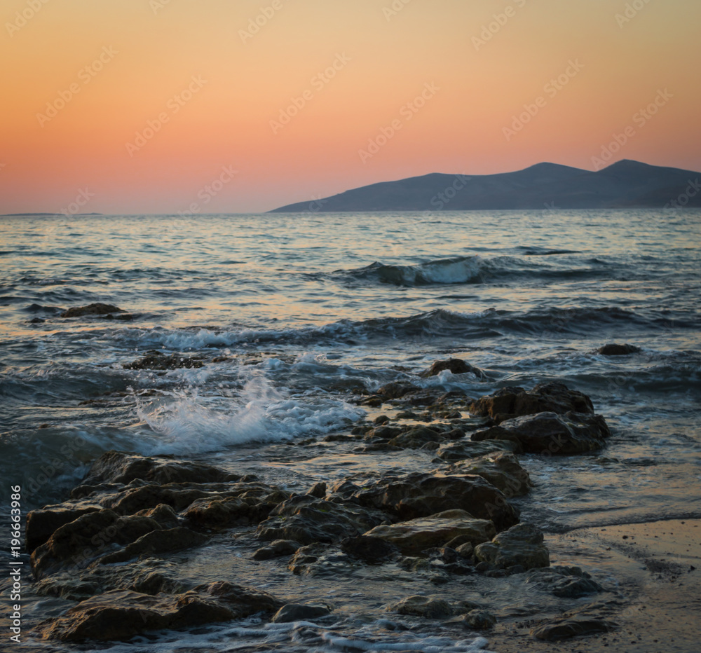 A long exposure of the sea as it breaks over the rocks and beach at golden hour, with the sun setting behind the mountains.