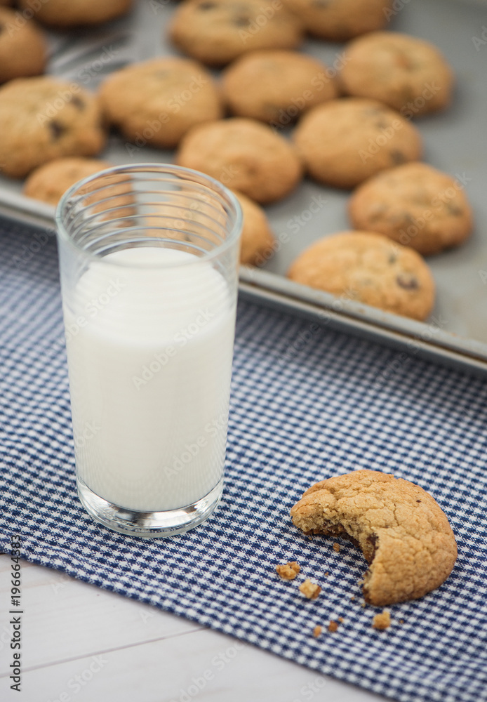 A glass of milk and a cookie with a bite taken out of it sit on a blue gingham napkin with a cookie sheet in the background A glass of milk and a cookie with a bite taken out of it sit on a blue gingham napkin with a cookie sheet in the background