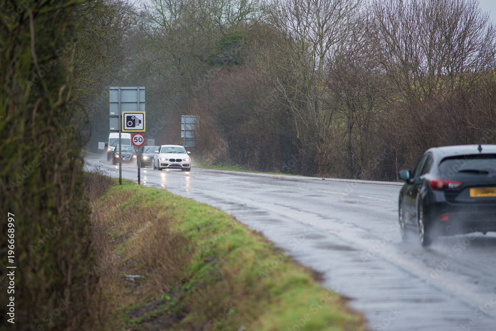 cars driving along water logged road with spray and rain and camera and 50 mph speed warning sign
