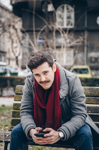 handsome hipster man with a vintage mustache using his smartphone outdoors. fashionable guy wearing a trendy coat texting his friends © mlasaimages