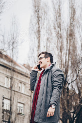 young attractive man talking on his smartphone outside. hipster guy with a mustache wearing a coat, eyelasses and a watch speaking on his mobile phone in the city