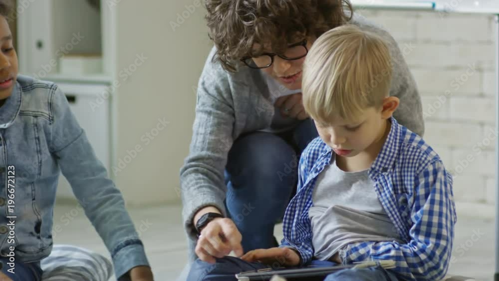 Female teacher explaining little boy how to use digital tablet while giving lesson to children in kindergarten Female teacher explaining little boy how to use digital tablet while giving lesson to children in kindergarten