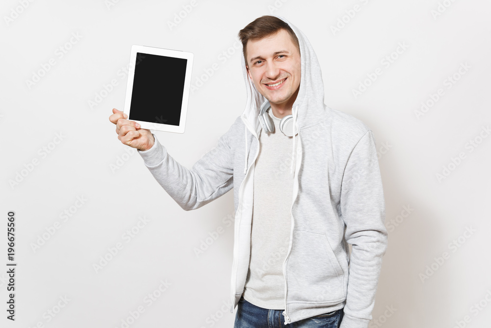 Young handsome smiling student in t-shirt and light sweatshirt with hood with headphones holds a tablet in hands and shows it to camera in studio on white background. Concept of technology Young handsome smiling student in t-shirt and light sweatshirt with hood with headphones holds a tablet in hands and shows it to camera in studio on white background. Concept of technology