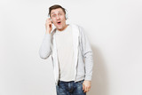 Young handsome student in t-shirt and light sweatshirt listening to music with white wireless headphones and puts hand to them in surprise in studio on white background. Concept of emotions