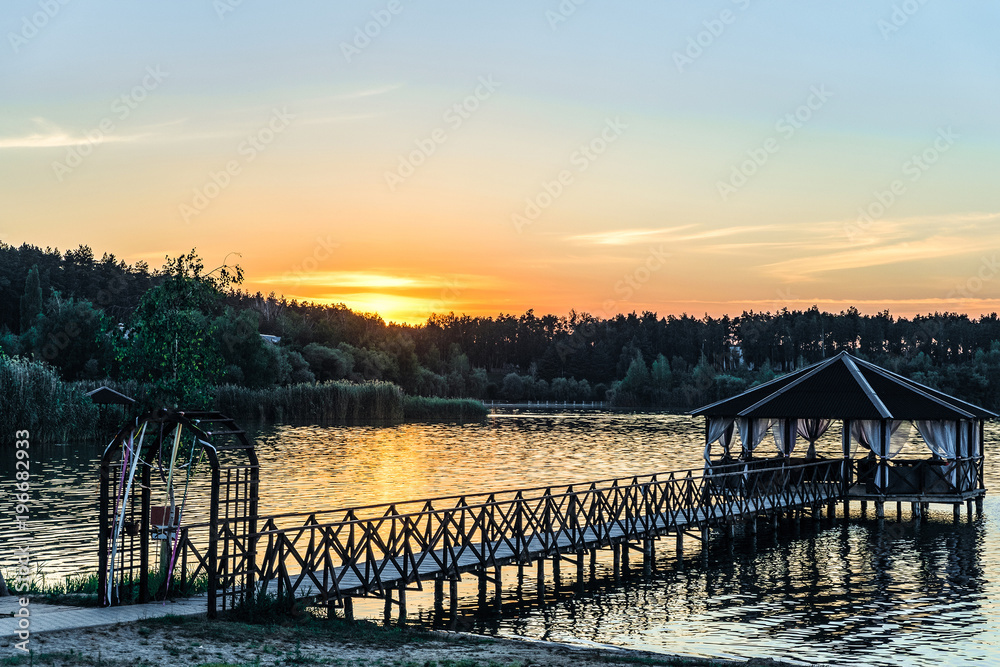Footbridge with a gazebo on the lake shore. Low evening sun at sunset.