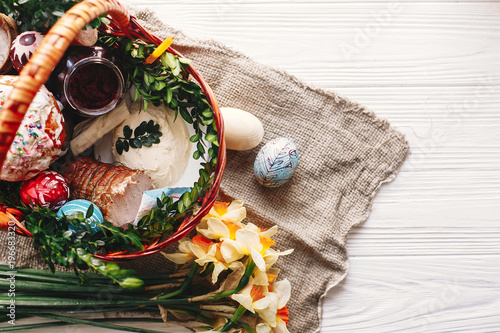 stylish basket with painted eggs, bread, ham,beets, butter on rustic wood background with spring flowers and candle, top view. easter food for blessing in church. happy Easter concept