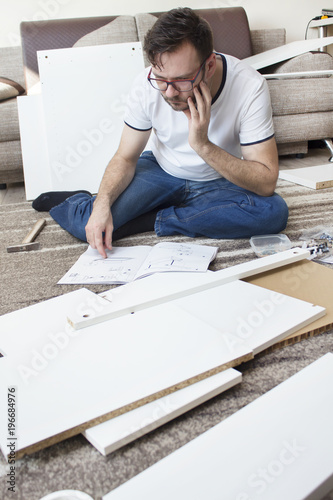 Brodaty mężczyzna w okularach, białej podkoszulce i jeansach siedzi na dywanie w zamyśleniu i czyta instrukcję montażu mebli. Dookoła niego leżą elementy do złożenia.