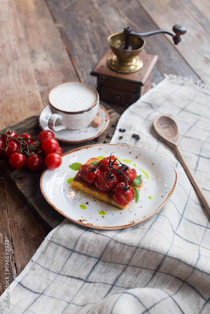 Traditional Italian vegetarian focaccia with fried cherry tomatoes, cappuccino cup with pieces of cane sugar and a hand grinder in the background, table covered with a tablecloth with a wooden spoon