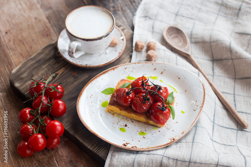Traditional Italian vegetarian focaccia with fried cherry tomatoes, cappuccino cup with pieces of cane sugar, table covered with a tablecloth with a wooden spoon close up