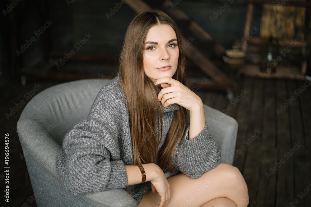 Dark haired sad woman in long grey knitted cozy sweater sitting in a chair at home near big window. Happy and Healthy girl with long beautiful legs with soft skin in studio. Daylight, serious teen. Dark haired sad woman in long grey knitted cozy sweater sitting in a chair at home near big window. Happy and Healthy girl with long beautiful legs with soft skin in studio. Daylight, serious teen.
