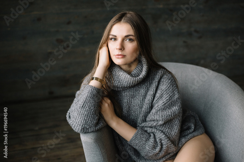 Dark haired sad woman in long grey knitted cozy sweater sitting in a chair at home near big window. Happy and Healthy girl with long beautiful legs with soft skin in studio. Daylight, serious teen.