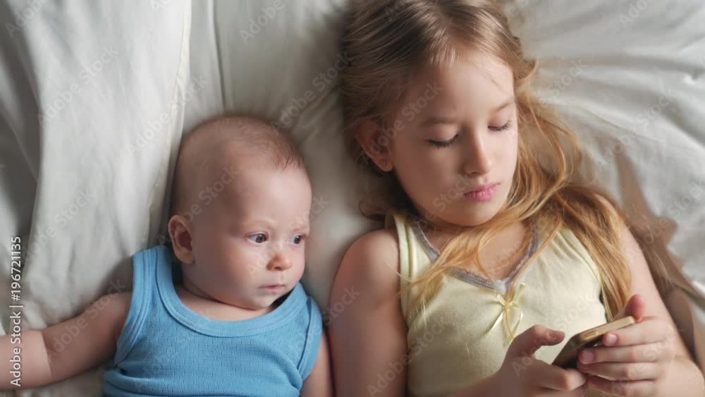 Little cute sister and baby brother is looking or playing with a smartphone on bed at home Little cute sister and baby brother is looking or playing with a smartphone on bed at home
