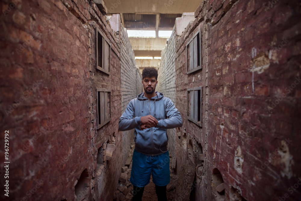 Portrait of focused motivated afro-american young handsome sportive man with earphones standing inside of the abandoned place in the middle of two walls and thinking.